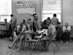 About a dozen partially-clothed residents unsupervised in a dayroom. Several sit on plastic chairs around a small table, on which sits another man with his knees pulled up to his chin. Others stand, looking out the window, or sit in chairs around the wall.