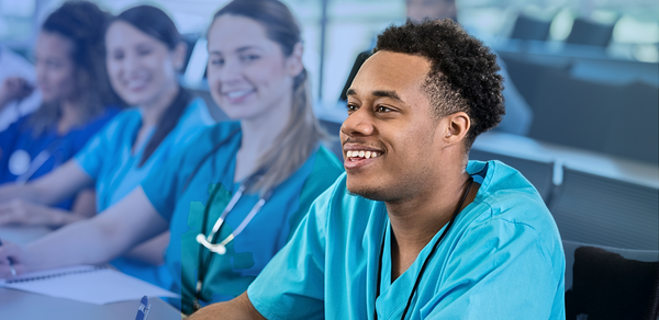 Young man in blue scrubs smiling in classroom with medical students beside him