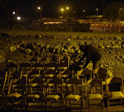 a shadowy figure leans over chairs arranged in rows in a rubble strewn nighttime outdoor setting.