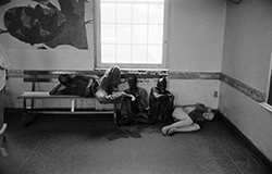 In the Dayroom of a Willowbrook ward, clothed and unclothed residents sit idle and bunched together on hard benches. One resident is curled up on the cold floor. This crowding and the lack of clothing or supervision led to the rapid spread of disease among residents.