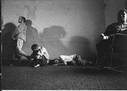 A stark image of residents lying on the floor, hugging their knees, leaning against the stained wall, and staring blankly into space. There are four people, but only one chair. The figures cast shadows on the wall, emphasizing their isolation. 