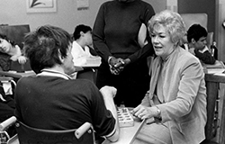 Elizabeth Connelly meets with a Staten Island Developmental Center program participant in 1985, in the center that was later named for her. Connelly sits knee-to-knee with a man in a wheelchair, their eyes meeting as they interact. Her hand extends toward his outreached hand. 