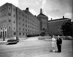 The iconic six-story hospital, Building 2 at Willowbrook State School, in the 1950s. Two horizontal wings extend from a narrow central tower topped with a lighted beacon. This building no longer exists; its site is now “The Great Lawn” of CSI/CUNY.