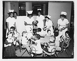 In a photograph intended to give a positive impression of conditions at Willowbrook, six nurses in starched white uniforms care for eleven well-dressed toddlers. The children sit in small chairs at tables with the nurses engaged with them.