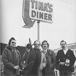 Geraldo Rivera and his ABC television news team in 1972. The group of men, holding cameras and microphones, stand beneath a neon sign that reads “Tina’s Diner” with a large arrow pointing downward.