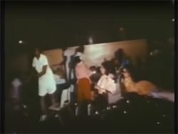 In a chaotic still-shot from the Rivera expose, a single nurse stands in a dark room illuminated by the camera light. A number of residents stand, sit, and lie around her as she works.