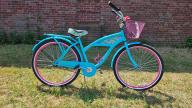 A Margaritaville branded, baby - blue - framed bike with black handleb ars, a pink basket, a cup holder, pedal breaks, and a black seat with pineapple - decals.