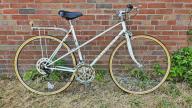 A white Peugeot branded bike with yellow rims, rear rack, gears and a handle break