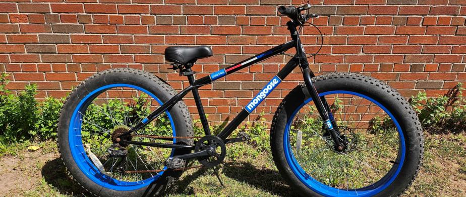 A Mongoose branded black and blue bike with large black wheels, black seat,  with handbreaks and gears