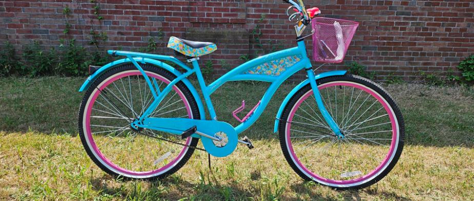 A Margaritaville branded, baby - blue - framed bike with black handleb ars, a pink basket, a cup holder, pedal breaks, and a black seat with pineapple - decals.