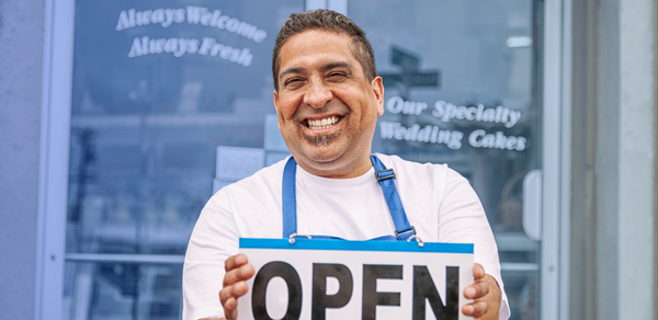 Man smiling in blue apron holding open sign in front of bakery doors