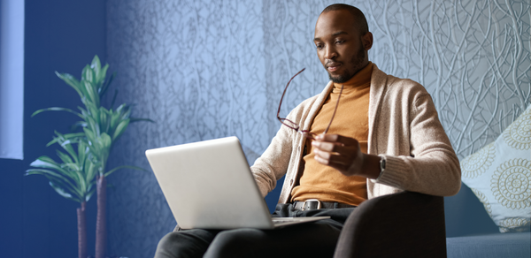 Man in tan turtleneck reviewing laptop in his lap while holding a pair of glasses