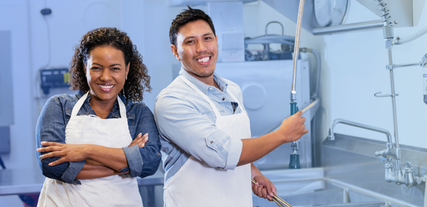 Man and woman smiling in commercial kitchen setting wearing white aprons