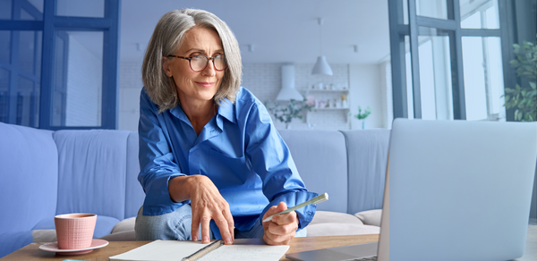 Older woman in blue shirt using laptop on sofa while taking notes