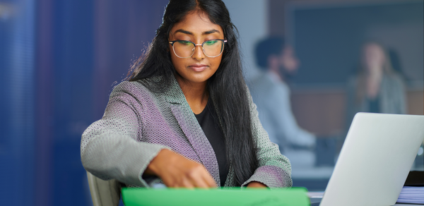 Young woman in suit and glasses searching through paper on desk with laptop