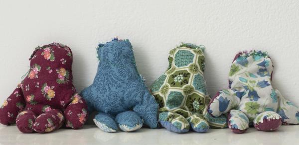 Photograph of 5 sculptures. Each sculpture is a small doll looking entity. Each is made of a different color fabric with patterns. 