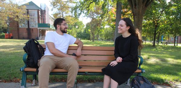 two students sitting on a wood bench surrounded by trees on a sunny day