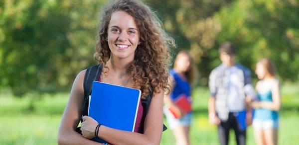 Student Standing In Field Holding Blue Book