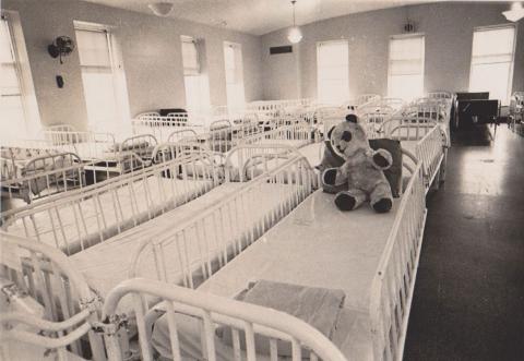 Dormitory, empty with tightly spaced beds, on one of which is a toy teddy bear