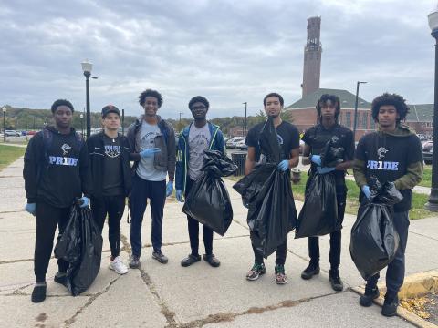 8 student athletes wearing their CSI uniform are standing in front 1 R Recreation building.​  They are holding black garbage bags filled with trash they picked up on campus.