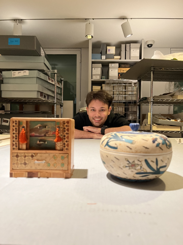 A student working in a collection storage room, posing with two small decorative objects: a wooden Japanese-style chest and a round lidded ceramic container featuring a floral motif. The Student is seen smiling and leaning on the table. A student working in a collection storage room, posing with two small decorative objects: a wooden Japanese-style chest and a round lidded ceramic container featuring a floral motif. The Student is seen smiling and leaning on the table.