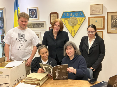 Five people gathered around a table, smiling, and looking at archival materials in a room with shelves, boxes, and a yellow-and-blue UNWLA crest on the wall. Five people gathered around a table, smiling, and looking at archival materials in a room with shelves, boxes, and a yellow-and-blue UNWLA crest on the wall.