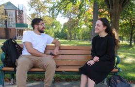 two students sitting on a wood bench surrounded by trees on a sunny day